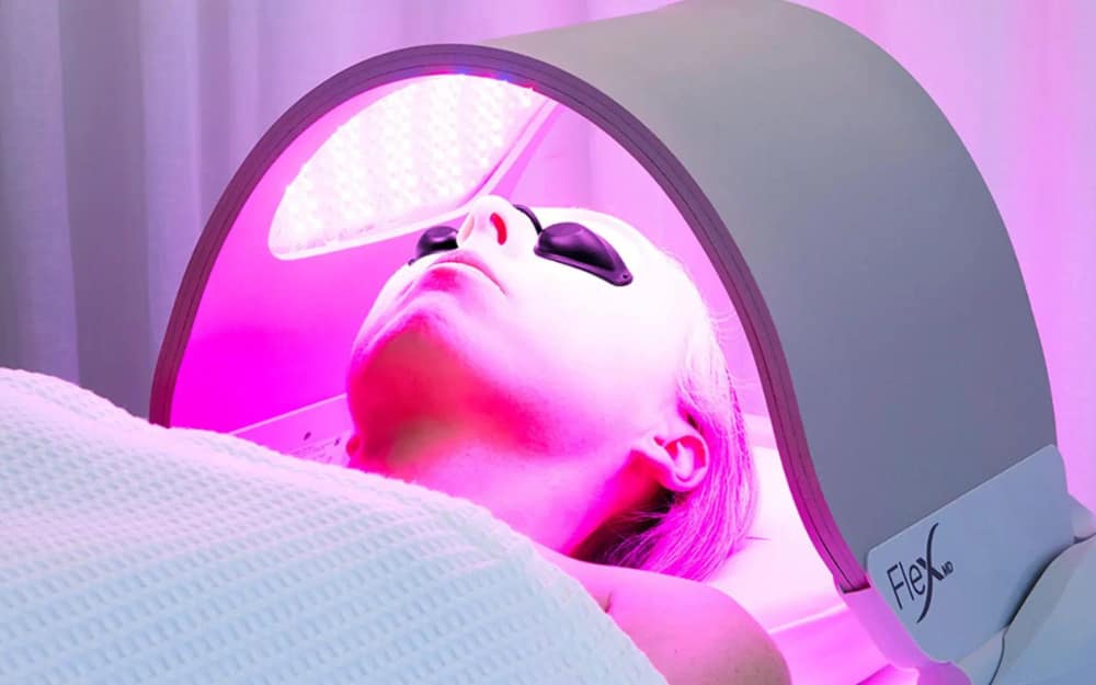 A close-up view of a patient lying down under a curved Dermalux LED phototherapy canopy emitting a combined blue and red light spectrum. The patient wears protective opaque goggles while the targeted panel is positioned over the face.