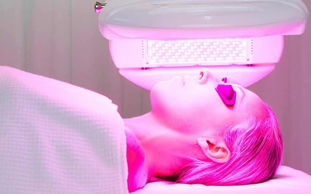 A clinical profile shot of a woman undergoing advanced phototherapy. A medical-grade LED panel arches over her face, flooding the skin with a highly concentrated, bright red light matrix.