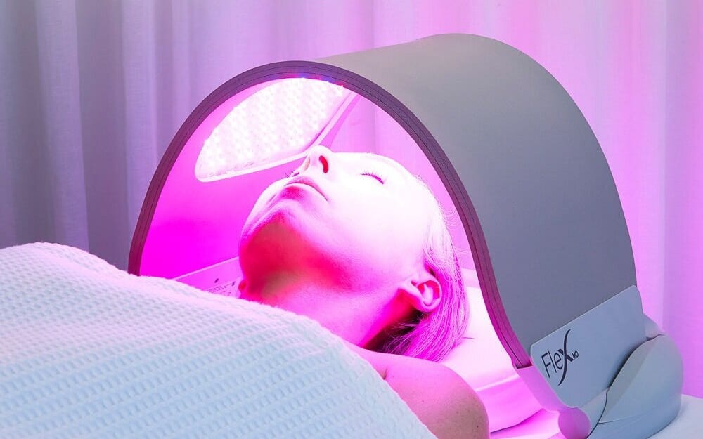 A patient resting comfortably on a clinical treatment bed during a post-treatment LED light therapy session. The overhead panel is active, casting a clean, warm therapeutic glow across the facial profile.